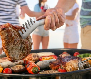 A person holds steak with metal tongs over a barbecue