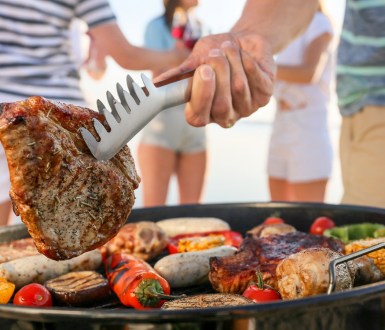 A person holds steak with metal tongs over a barbecue