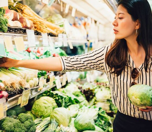 A young asian woman reaches for some vegetables in a supermarket. She is holding a lettuce.