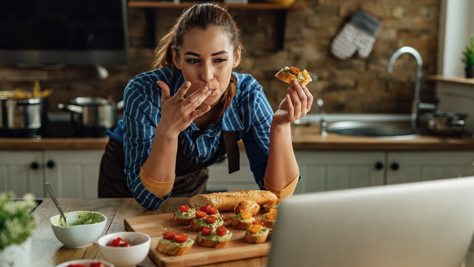 A woman eats avocado and tomato on bread while looking at a laptop