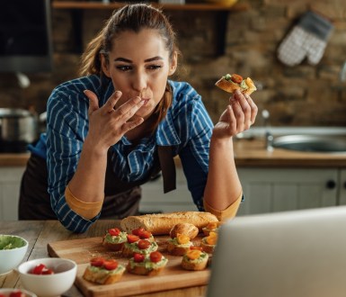 A woman eats avocado and tomato on bread while looking at a laptop