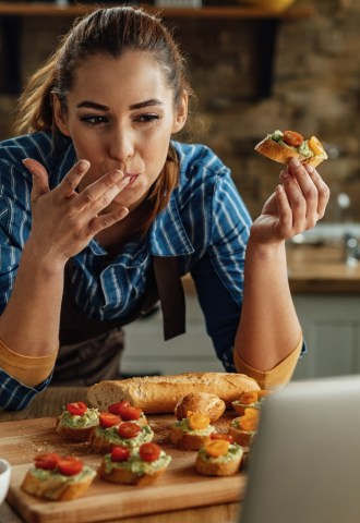 A woman eats avocado and tomato on bread while looking at a laptop