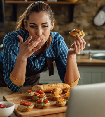 A woman eats avocado and tomato on bread while looking at a laptop